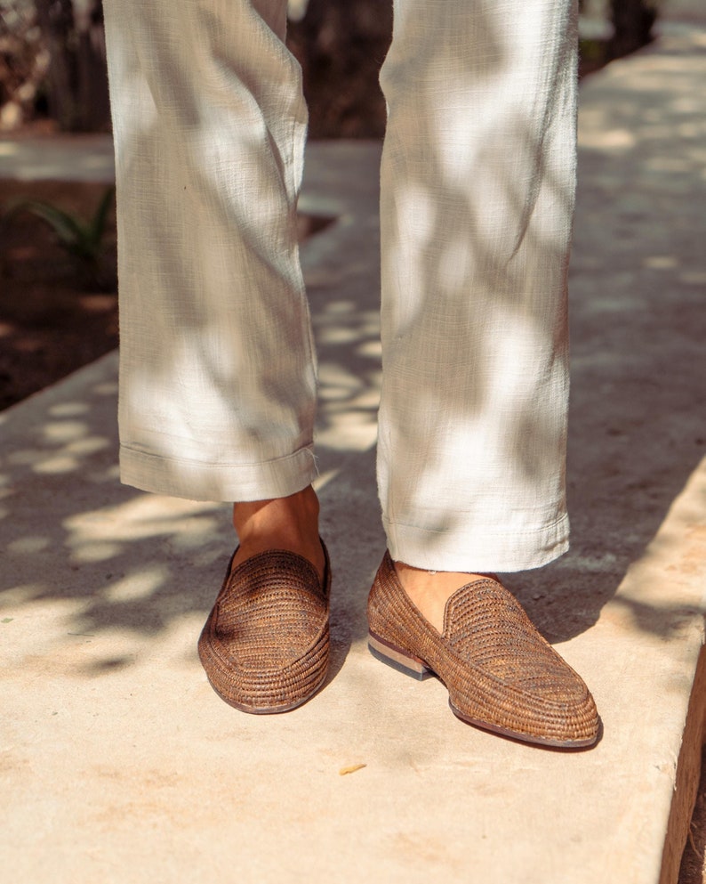 K&ouml;nnte beinhalten: Ein Paar braune geflochtene Lederschuhe mit einer hellbraunen Sohle. Die Schuhe werden von einer Person getragen, die eine wei&szlig;e Hose tr&auml;gt.
