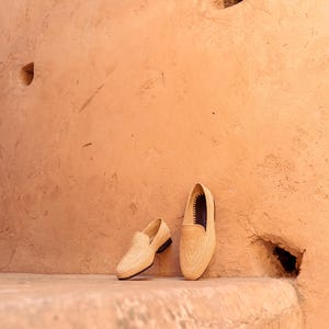 May include: Two beige woven loafers with black soles are displayed against a textured, peach-colored wall. The shoes are positioned on a matching ledge, showcasing their intricate weave and classic design. The loafers are a stylish footwear option.