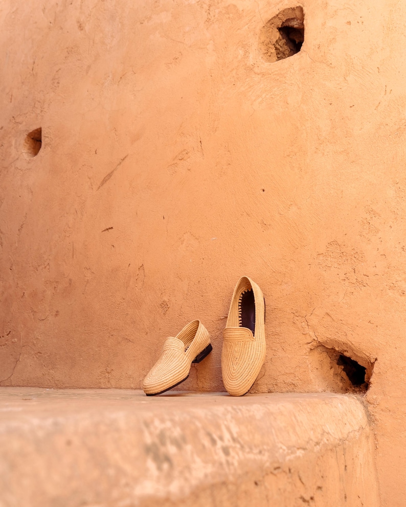 May include: Two beige woven loafers with black soles are displayed against a textured, peach-colored wall. The shoes are positioned on a matching ledge, showcasing their intricate weave and classic design. The loafers are a stylish footwear option.