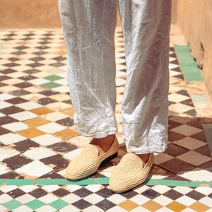 May include: A pair of beige woven loafers with a rounded toe. The shoes are worn with light-colored linen pants. The shoes are on a tiled floor with a geometric pattern of black, white, green, and yellow tiles.