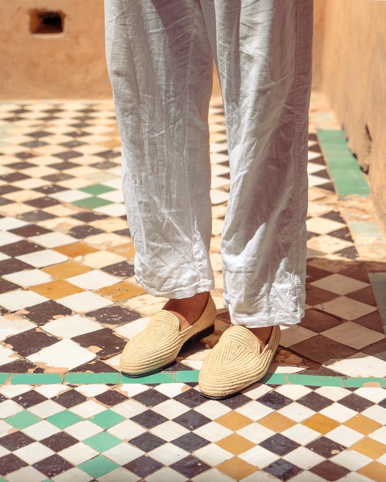 May include: A pair of beige woven loafers with a rounded toe. The shoes are worn with light-colored linen pants. The shoes are on a tiled floor with a geometric pattern of black, white, green, and yellow tiles.