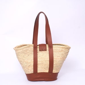 Peut inclure: Un grand sac fourre-tout en paille tress&eacute;e avec des anses et des accents en cuir marron. Le sac a un haut large et ouvert et un fond arrondi.