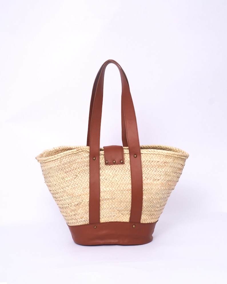 Peut inclure: Un grand sac fourre-tout en paille tress&eacute;e avec des anses et des accents en cuir marron. Le sac a un haut large et ouvert et un fond arrondi.