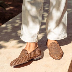 K&ouml;nnte beinhalten: Ein Paar braune geflochtene Lederschuhe mit leicht spitzer Zehenpartie. Die Schuhe werden von einer Person getragen, die eine wei&szlig;e Hose tr&auml;gt.