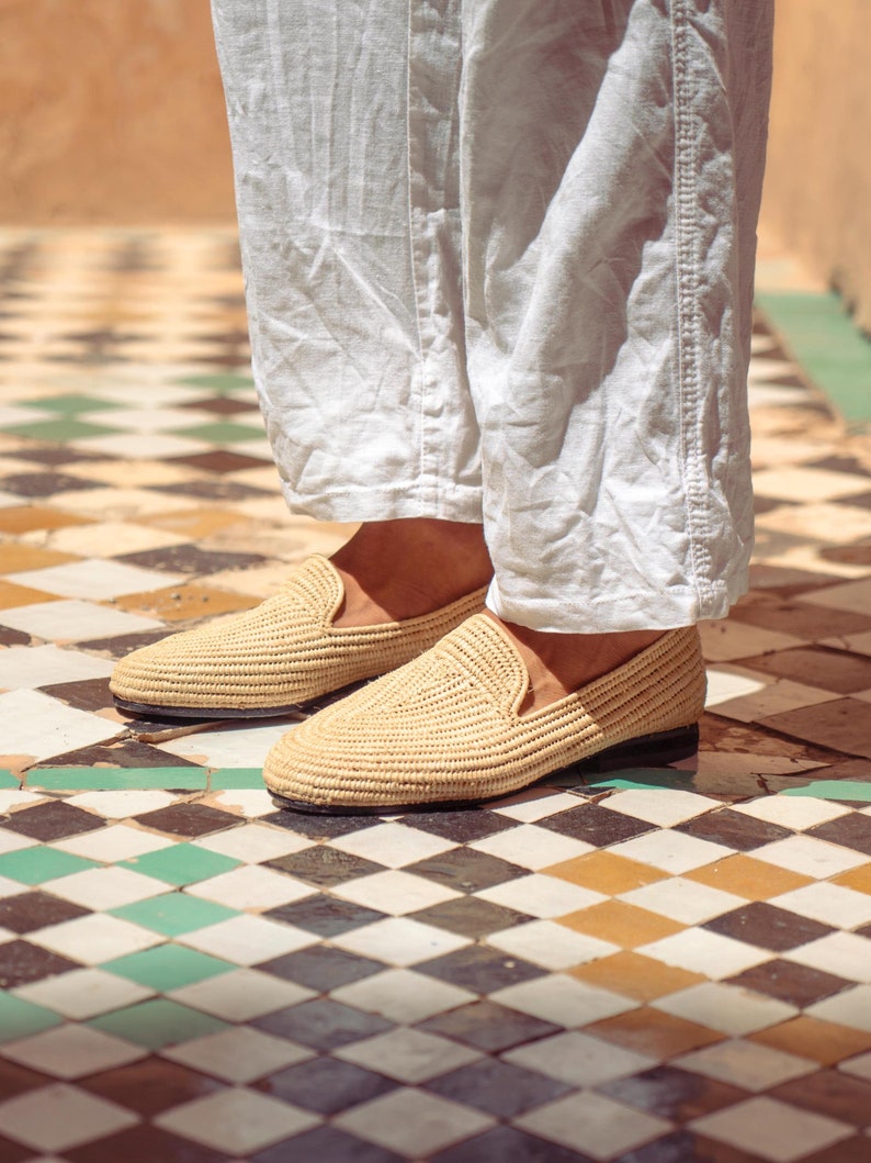 May include: Close-up of woven raffia loafers in a natural beige color. The shoes have a classic slip-on design with a dark sole. The image shows the shoes worn with white linen pants, set against a patterned tile floor.