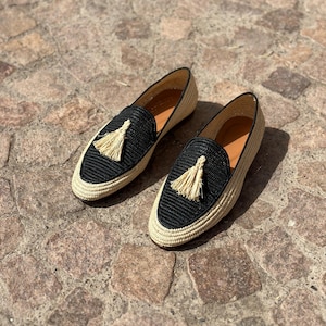 Puede incluir: Un par de mocasines tejidos negros y beige con un detalle de borla. Los mocasines tienen una suela plana y una punta puntiaguda.