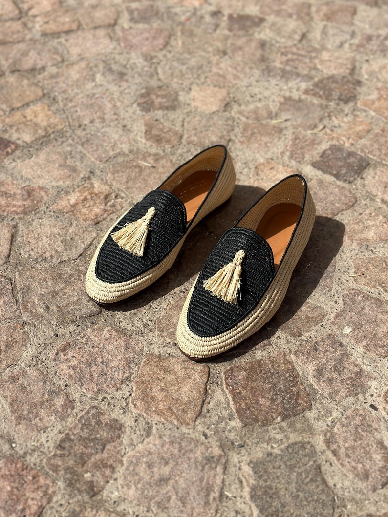 Puede incluir: Un par de mocasines tejidos negros y beige con un detalle de borla. Los mocasines tienen una suela plana y una punta puntiaguda.