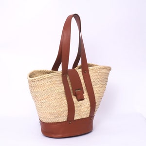 Peut inclure: Un grand sac cabas en paille avec des anses et des finitions en cuir marron. Le sac a une texture tiss&eacute;e et un fond en cuir marron.