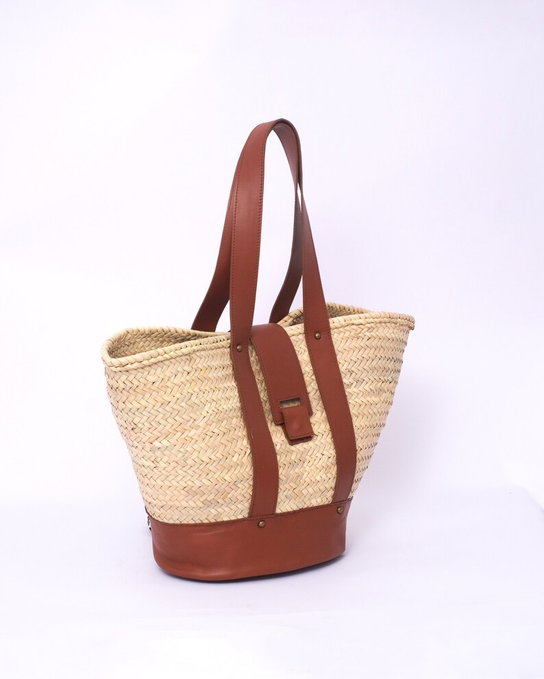 Peut inclure: Un grand sac cabas en paille avec des anses et des finitions en cuir marron. Le sac a une texture tiss&eacute;e et un fond en cuir marron.