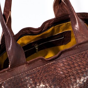 Puede incluir: Un bolso tote de cuero marr&oacute;n con un patr&oacute;n tejido. El bolso tiene un bolsillo con cremallera en el interior y un forro amarillo.