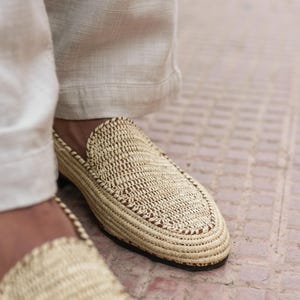 Puede incluir: Primer plano de un par de zapatillas de paja tejidas con ribete marr&oacute;n. Las zapatillas las lleva una persona que lleva pantalones blancos.
