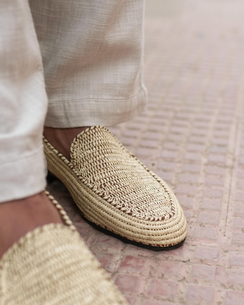 Puede incluir: Primer plano de un par de zapatillas de paja tejidas con ribete marr&oacute;n. Las zapatillas las lleva una persona que lleva pantalones blancos.