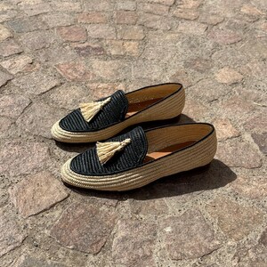 Puede incluir: Un par de mocasines de borlas tejidos en negro y beige. Los mocasines tienen una parte superior tejida con un detalle de borla negra. Los mocasines est&aacute;n sobre una superficie de piedra.
