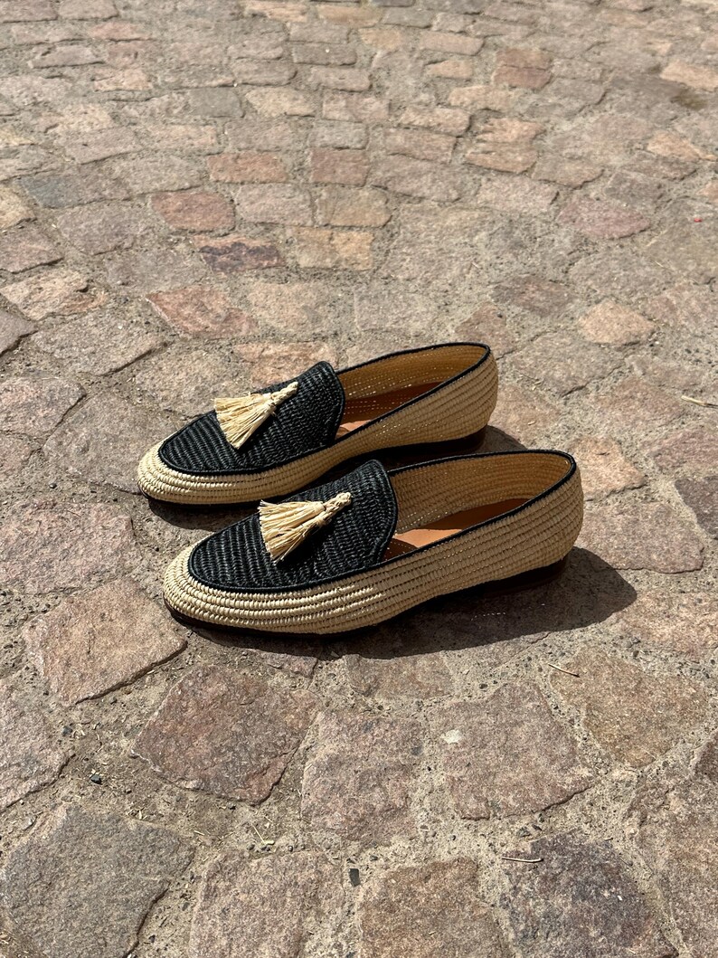 Puede incluir: Un par de mocasines de borlas tejidos en negro y beige. Los mocasines tienen una parte superior tejida con un detalle de borla negra. Los mocasines est&aacute;n sobre una superficie de piedra.