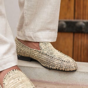 Pode incluir: Um par de mocassins de palha tran&ccedil;ada com acabamento preto. Os mocassins s&atilde;o usados por uma pessoa que usa cal&ccedil;as brancas.
