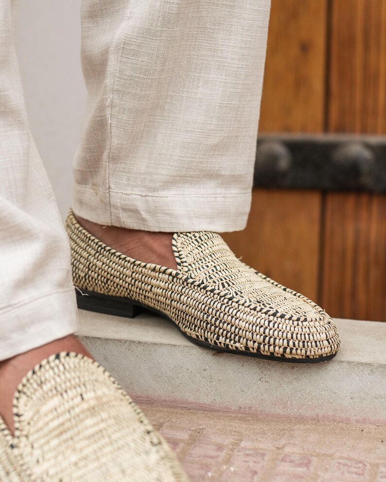 Pode incluir: Um par de mocassins de palha tran&ccedil;ada com acabamento preto. Os mocassins s&atilde;o usados por uma pessoa que usa cal&ccedil;as brancas.
