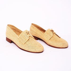 Può includere: Un paio di mocassini in pelle intrecciata beige con lacci. Le scarpe hanno una punta arrotondata e un tacco basso.