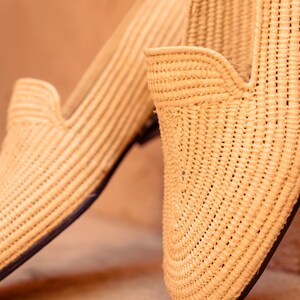 May include: Close-up of two woven raffia loafers. The shoes are a natural beige color with a textured, striped pattern. The soles are dark, and the design features a low-cut vamp. The shoes are likely for casual wear.