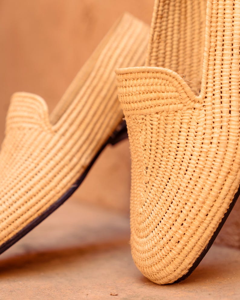 May include: Close-up of two woven raffia loafers. The shoes are a natural beige color with a textured, striped pattern. The soles are dark, and the design features a low-cut vamp. The shoes are likely for casual wear.
