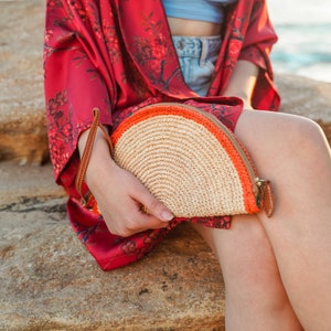 May include: A half-circle straw bag with a red and orange trim. The bag has a zipper closure and a brown leather strap.