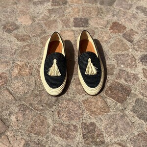 Puede incluir: Un par de mocasines de borlas tejidos en negro y beige. Los mocasines tienen una parte superior tejida de paja y un forro de cuero negro. Las borlas son de paja beige.