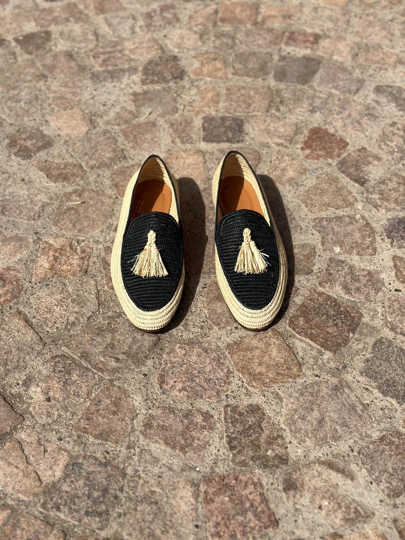 Puede incluir: Un par de mocasines de borlas tejidos en negro y beige. Los mocasines tienen una parte superior tejida de paja y un forro de cuero negro. Las borlas son de paja beige.