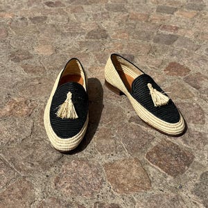 Puede incluir: Un par de mocasines de borlas tejidos en negro y beige. Los mocasines tienen una parte superior tejida en negro con un ribete tejido en beige y una borla beige.