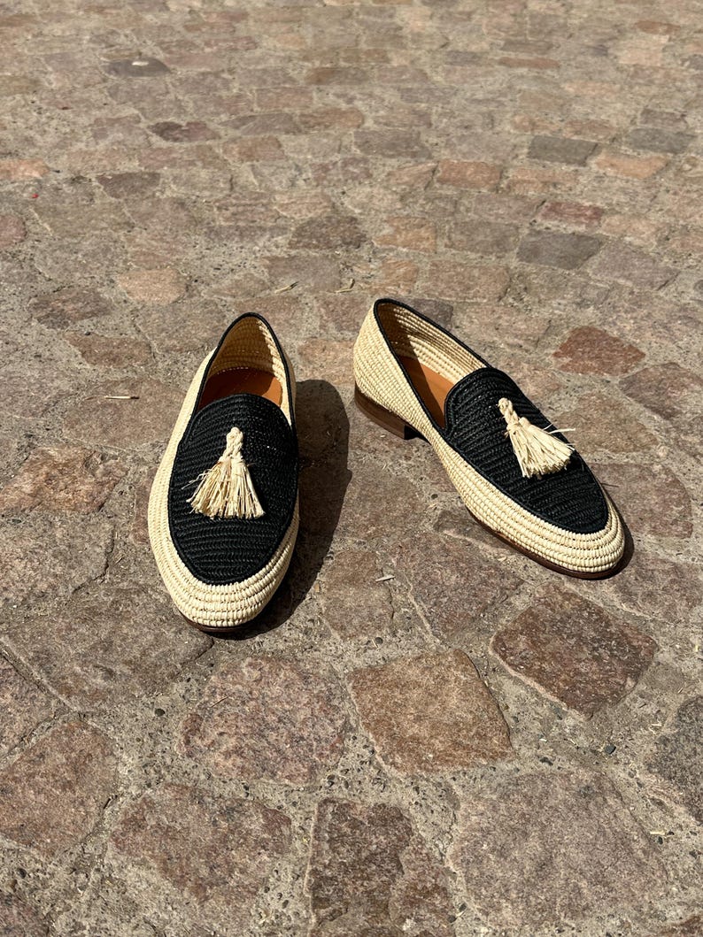 Puede incluir: Un par de mocasines de borlas tejidos en negro y beige. Los mocasines tienen una parte superior tejida en negro con un ribete tejido en beige y una borla beige.