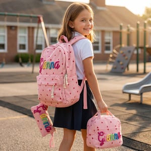 Op de afbeelding: Een roze rugzak, een bijpassend etui en een lunchtas, allemaal voorzien van een rood aardbeienpatroon en de naam Gianna geborduurd in kleurrijke, verhoogde letters. Elk item heeft witte touw-ritsen en een gestructureerd stoffen ontwerp.