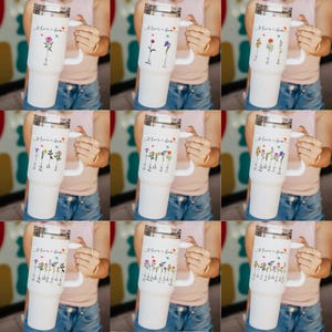 Puede incluir: Nueve vasos de acero inoxidable blanco con asa negra y dise&ntilde;o floral. Cada vaso tiene el texto "Mom's Garden" y los nombres de diferentes flores.