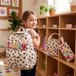 Può includere: Un set composto da zaino, borsa per il pranzo e astuccio in velluto a coste color crema con motivo a margherite nere. Ogni articolo è personalizzato con il nome Lainey in lettere di filato colorato e testurizzato e un piccolo fiore in tessuto rosa.