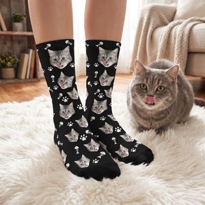 May include: Black crew socks featuring a repeating pattern of a grey tabby cat face with its tongue out, interspersed with white fish bone skeletons and paw print icons. The socks have a mid-calf length and a consistent graphic print design.