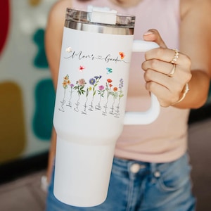 Puede incluir: Taza de viaje de acero inoxidable blanca con asa negra. La taza tiene un dise&ntilde;o floral con el texto "Mom's Garden" y una lista de nombres debajo de las flores.
