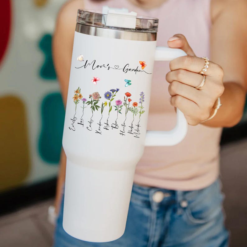 Puede incluir: Taza de viaje de acero inoxidable blanca con asa negra. La taza tiene un dise&ntilde;o floral con el texto "Mom's Garden" y una lista de nombres debajo de las flores.