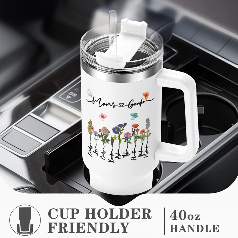 Puede incluir: Un vaso de acero inoxidable blanco de 40 onzas con tapa y asa. El vaso tiene un dise&ntilde;o floral con el texto "Mom's Garden" y un coraz&oacute;n. El vaso est&aacute; en un portavasos de coche.