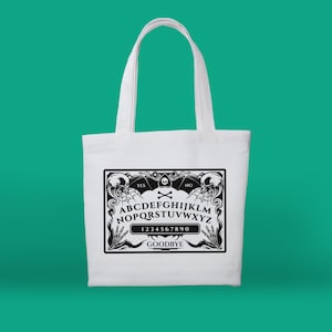 May include: White canvas tote bag with a black and white graphic of a Ouija board. The board features the letters A-Z, numbers 0-9, and the words "YES", "NO", and "GOODBYE".