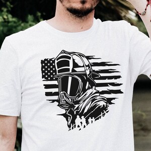 May include: White t-shirt featuring a black and white graphic of a firefighter wearing a helmet and gas mask in front of an American flag.