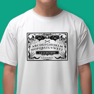 May include: White t-shirt with a black and white graphic of a Ouija board. The board features the letters A-Z, numbers 0-9, and the words "YES", "NO", and "GOODBYE".