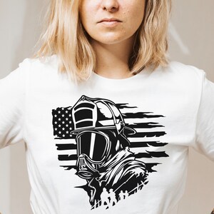 May include: A white t-shirt featuring a black and white graphic of a firefighter wearing a helmet and mask in front of an American flag.