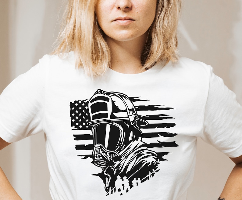 May include: A white t-shirt featuring a black and white graphic of a firefighter wearing a helmet and mask in front of an American flag.