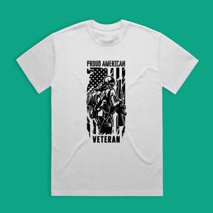 May include: White t-shirt with a black and white graphic of a soldier standing in front of an American flag. The text "Proud American Veteran" is printed on the shirt.