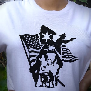 K&ouml;nnte beinhalten: Schwarz-wei&szlig;es Silhouettendesign auf einem wei&szlig;en T-Shirt mit einem Polizisten, einem Hubschrauber, einem Soldaten, Handschellen und einer amerikanischen Flagge.