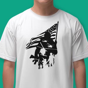 May include: A white t-shirt with a black silhouette of a soldier holding an American flag. The soldier is standing in front of a group of other soldiers, and there are helicopters flying overhead.