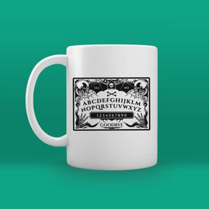May include: White ceramic mug with a black and white image of a Ouija board. The board has the letters A-Z, numbers 0-9, and the words "YES", "NO", and "GOODBYE".