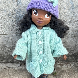 Peut inclure: Une poupée aux cheveux noirs bouclés portant un bonnet en tricot violet clair avec un petit nœud vert clair, et un manteau en tricot vert clair avec un col, quatre boutons blancs sur le devant et deux petites poches, avec des chaussures noires.