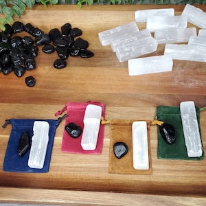 May include: Four small velvet pouches each containing a selenite crystal wand and a black obsidian stone. The pouches are red, blue, brown, and green. There are also several selenite crystal wands on a wooden surface.