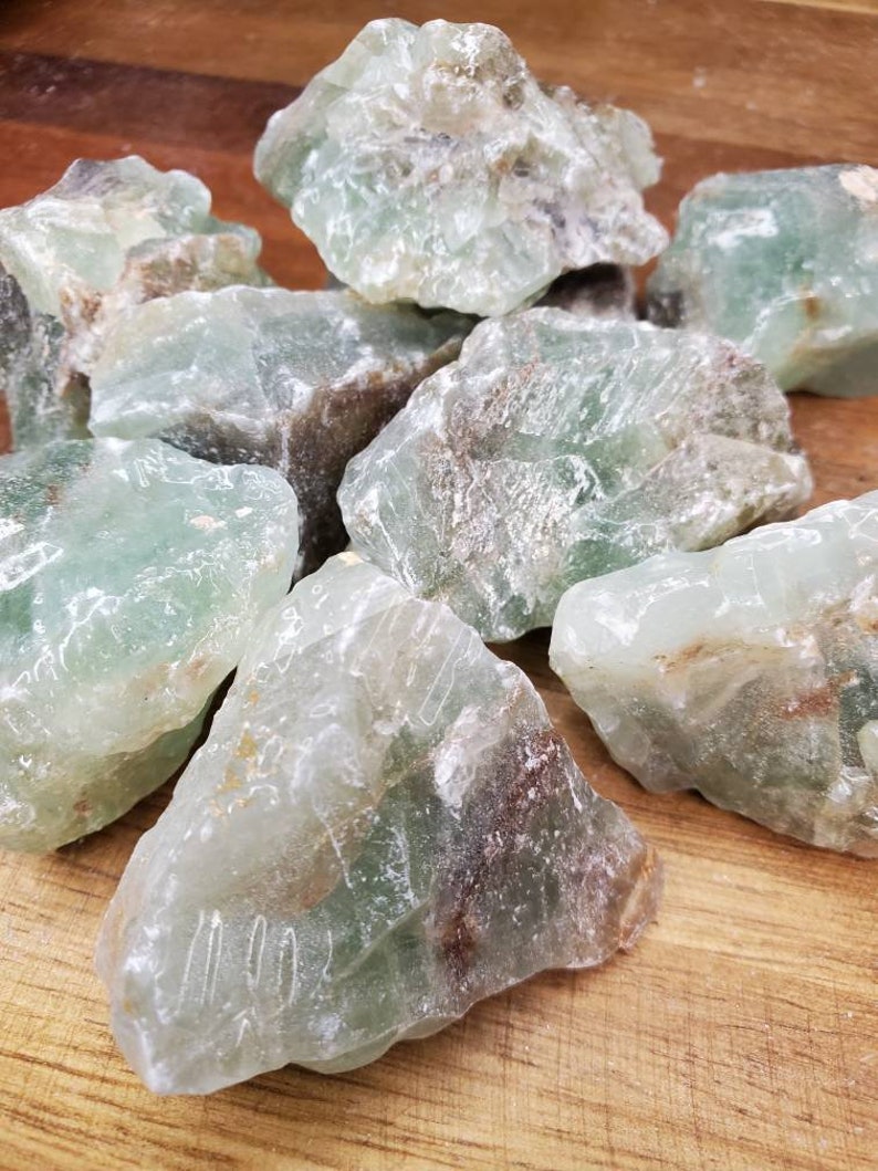 May include: A collection of rough, green fluorite crystals with brown inclusions. The crystals are various sizes and shapes, and have a translucent appearance.