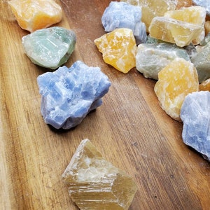 May include: A collection of raw, natural crystals in various shades of blue, green, and yellow. The crystals are arranged on a wooden surface.