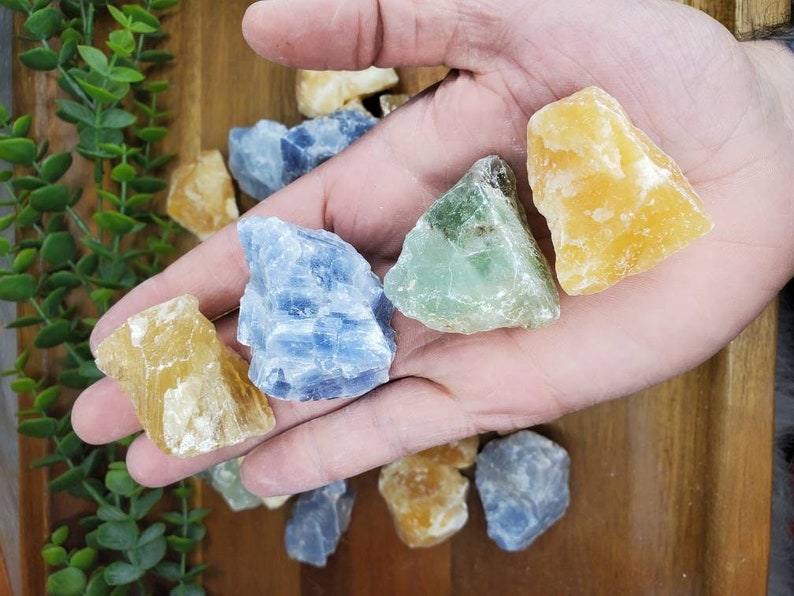 May include: A collection of six rough, natural crystals in various colors, including blue, green, and yellow. The crystals are arranged in a hand, showcasing their unique shapes and textures.