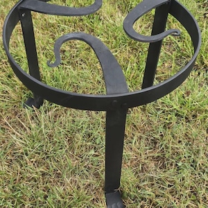 May include: A black metal stand with a circular top and three curved supports. The stand has three legs and is designed to hold a pot or other item. The stand is sitting on grass.